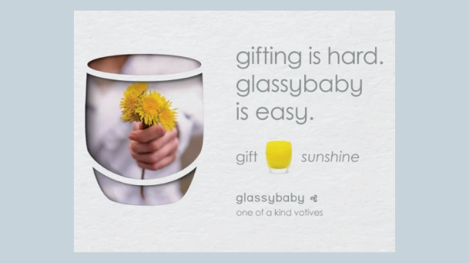 A hand holding dandelions within a Glassybaby candle outline. Text reads 'gifting is hard. Glassybaby is easy.' and 'one of a kind votives' next to the Glassybaby logo.