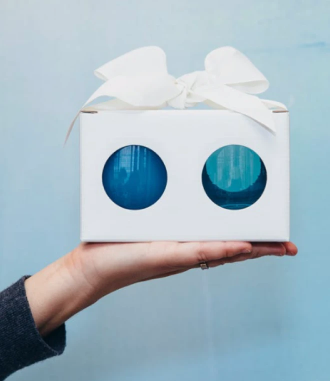 A hand holding a white gift box with a white bow on top and two circular windows revealing blue glass items inside.