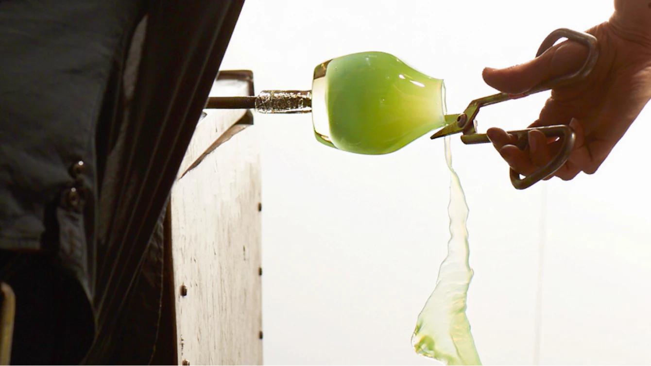 Close-up of a glass artist shaping a molten green glass piece with metal scissors.