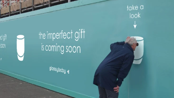 Man peering through a peephole on a wall featuring the Glassybaby logo, text 'the imperfect gift is coming soon' with the phrase 'take a look' pointing towards the peephole.