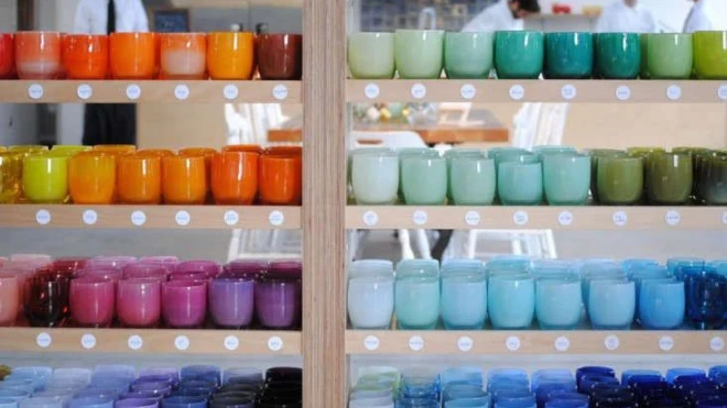 Multiple shelves displaying a colorful array of Glassybaby candles in a variety of hues, from vibrant oranges and reds to calming blues and greens, sorted by color.