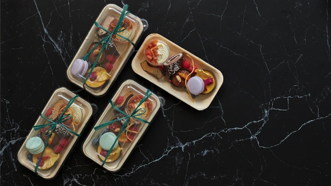 Top-down view of four elegantly packaged gourmet desserts in eco-friendly containers placed on a black marble surface.