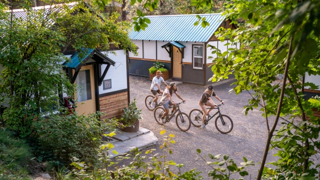 Four people ride bikes on a gravel path between cabins with blue metal roofs, surrounded by green trees