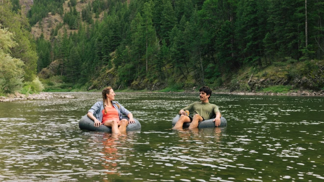 Two people relaxing on inner tubes in a calm river surrounded by dense forest and green hills