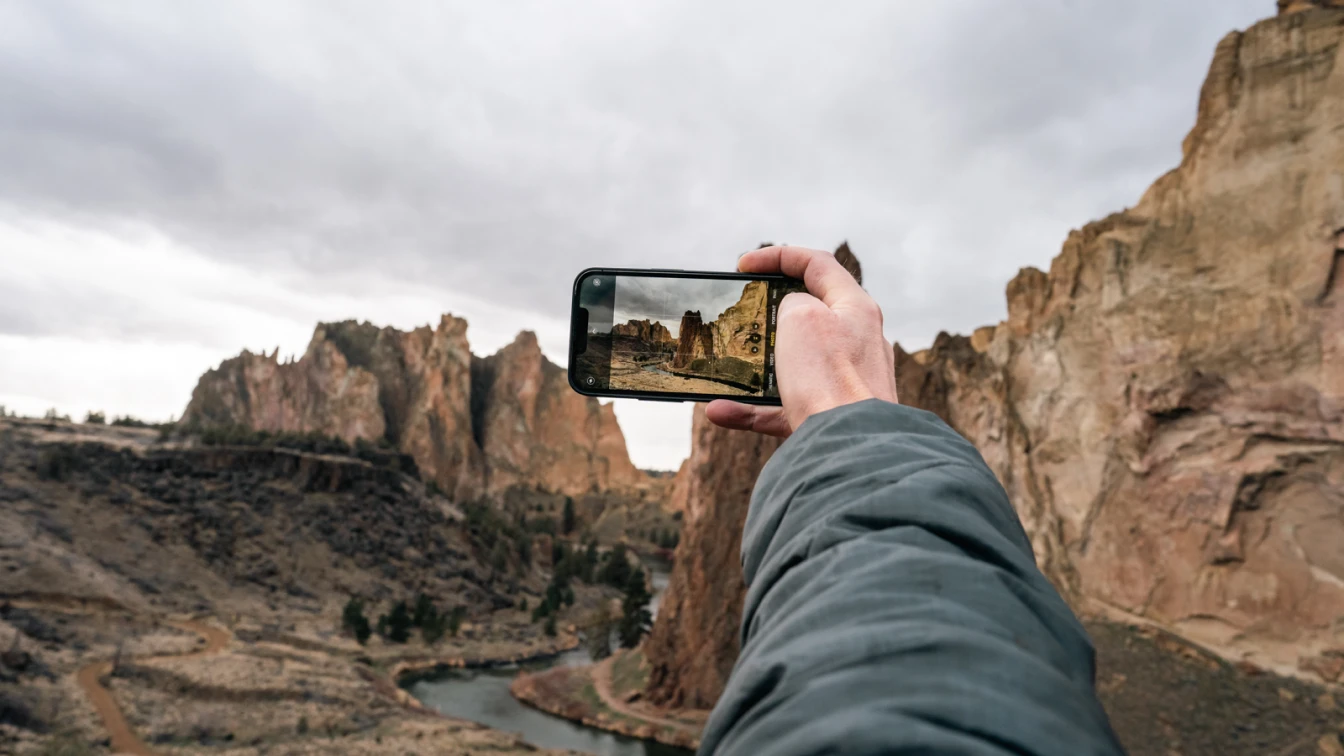 A person holds a smartphone to capture a photo of a stunning rocky landscape with rugged cliffs and a winding river below, under a cloudy sky, perfect for sharing on social media.