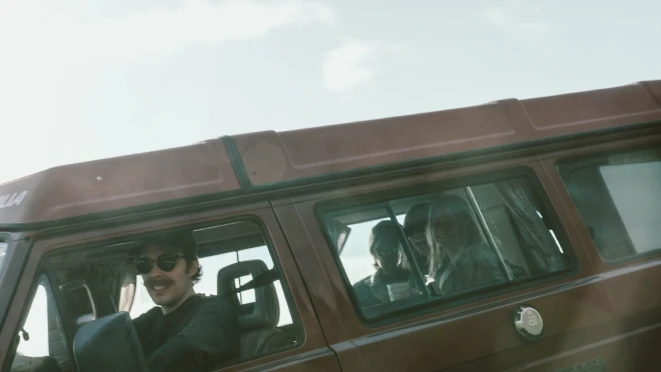 A smiling man wearing sunglasses and a cap drives a vintage brown van with passengers in the backseat. The van has a retro look, adding to the nostalgic feel of the image.