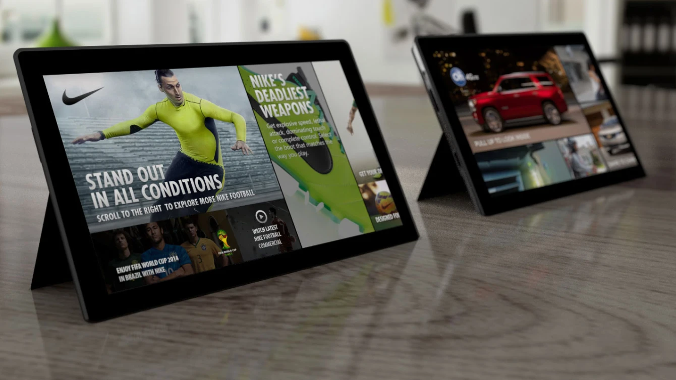 Close-up of two tablet devices on a table. One tablet shows a Nike ad with a female athlete and the text 'Stand out in all conditions.' The other tablet shows an ad for a red car.