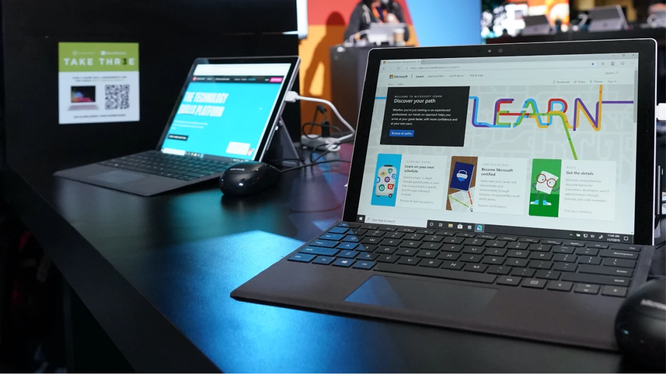 Two laptops displaying Microsoft Learn content are set up on a dark table at an event. The closest laptop features the Microsoft Learn homepage with the text "Discover your path.