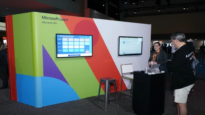 Two people interacting at a Microsoft Learn Event booth that highlights a security administrator game board and a learning path interface.