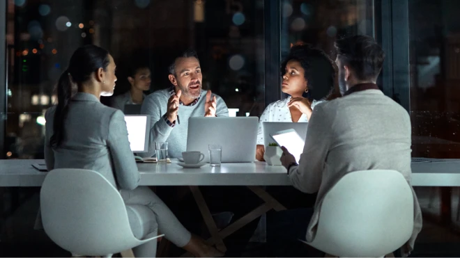 A diverse group of professionals engaged in a discussion during a nighttime meeting at a table with laptops and tablets and a cityscape through windows in the background.