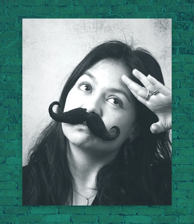 A black and white picture of a woman posing playfully with long dark hair wearing an fake mustache against a green brick wall. 