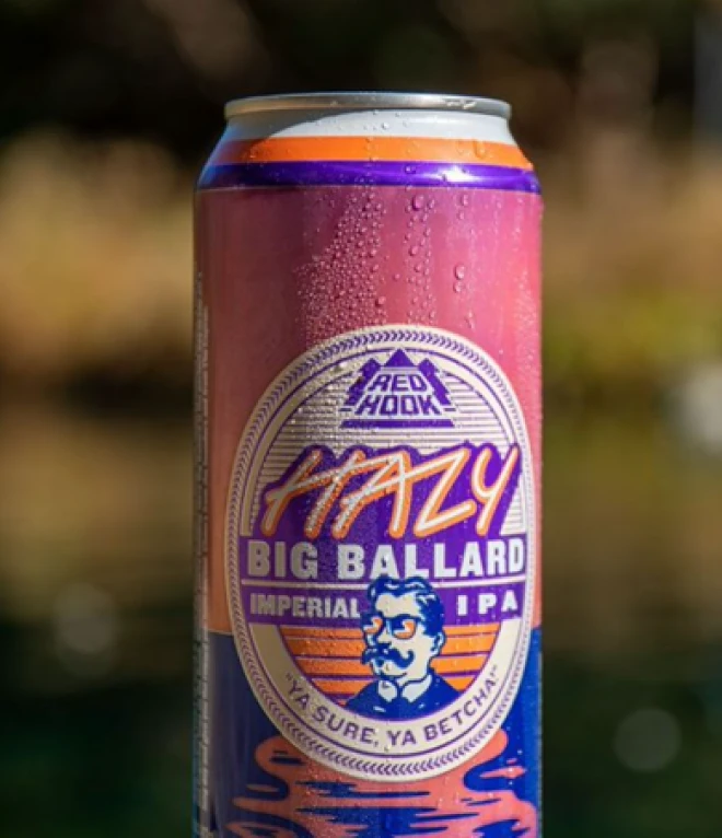 Close-up of a Red Hook Hazy Big Ballard Imperial IPA can with a pink and purple gradient background, condensation droplets, and a logo featuring a bearded man. The can's label reads "Hazy Big Ballard Imperial IPA" and includes the tagline "Ya Sure, Ya Betcha!"