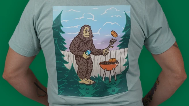 A close-up image of the back of a person wearing a light gray T-shirt featuring a graphic depicting a friendly sasquatch grilling burgers in a backyard setting and holding a Red Hook beer can.