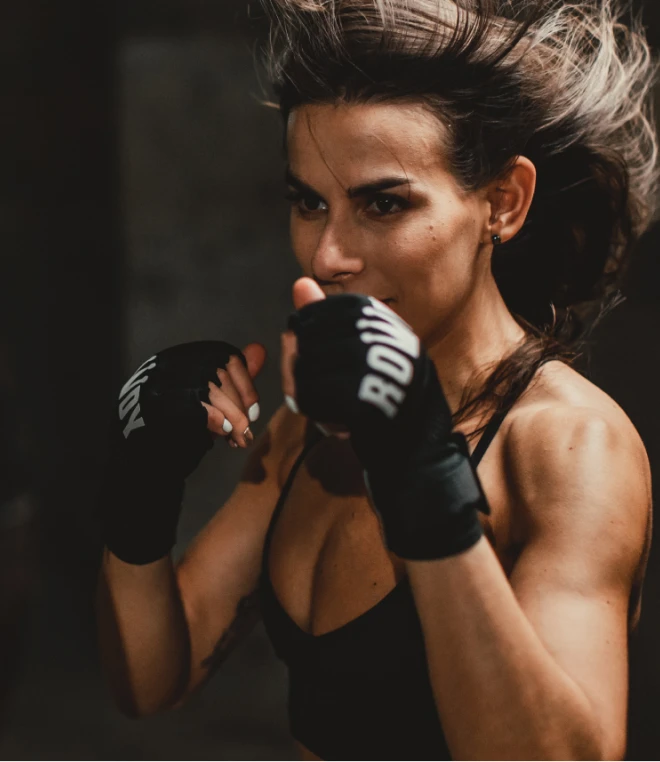 A woman against a dark background is wearing boxing gloves labeled "Rowdy" and in a boxing stance.