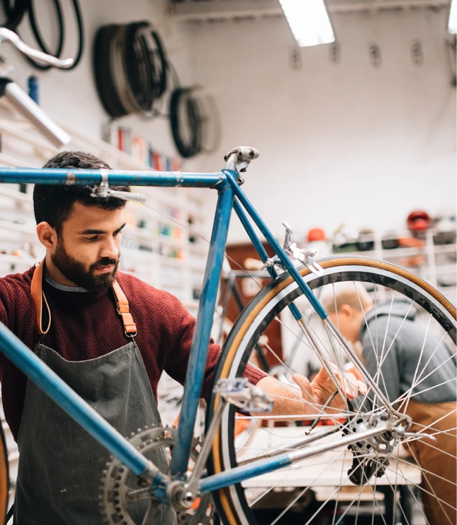 A bearded man wearing an apron is in a bicycle workshop, focused on repairing a blue bicycle.