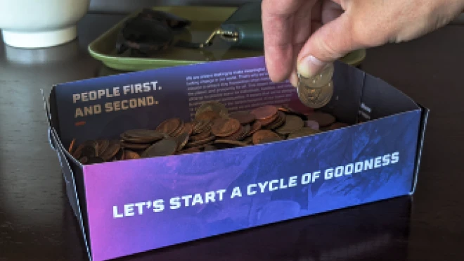 A hand placing coins into a makeshift coin tray made from a folded Verity Credit Union promotional mailer. The mailer has the text "Let's Start a Cycle of Goodness" visible on the side.