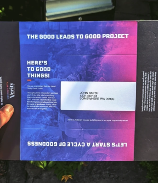 A promotional mailer from Verity Credit Union, held in a person's hand. The mailer has the text "The Good Leads to Good Project" and "Here's to Good Things!"